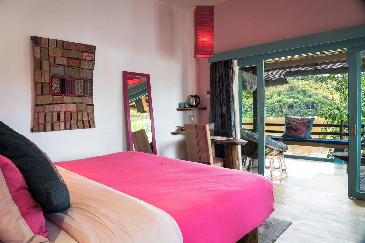 A bedroom with a large bed, a mirror, a desk, and a view of a garden through an open door.