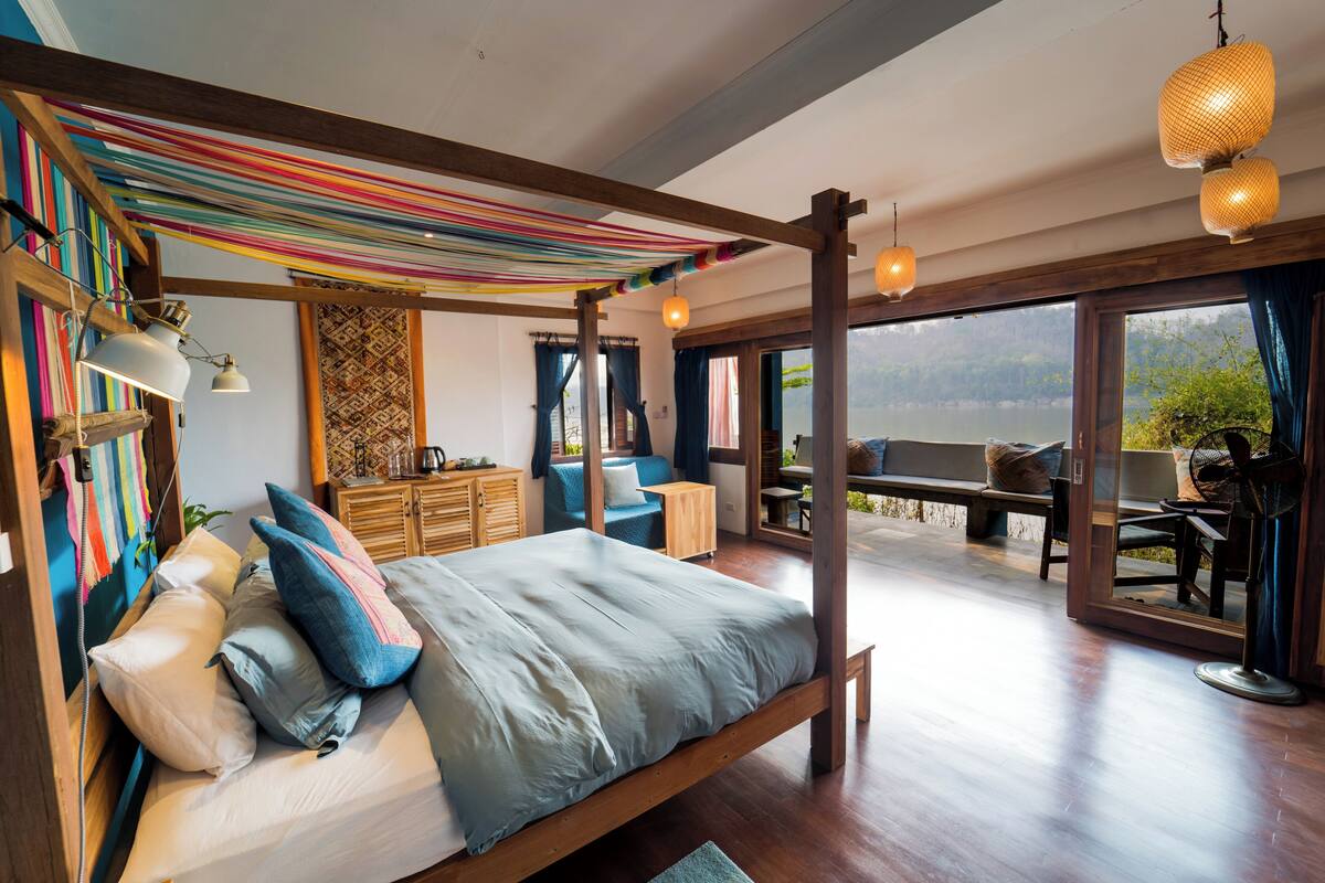 A spacious bedroom with a large bed, wooden headboard, and a view of a lake through large windows.