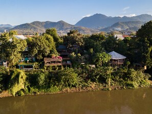A riverside view of a resort with multiple wooden structures, including a large building with a thatched roof and several smaller huts. - Ock Pop Tok Mekong Villa (Luang Prabang)