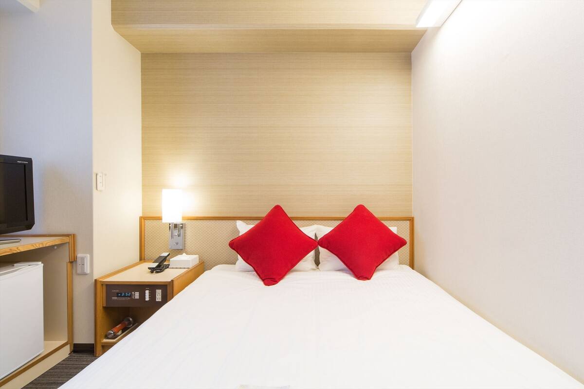 A hotel room with a bed, a nightstand, a television, and a wall-mounted lamp.
