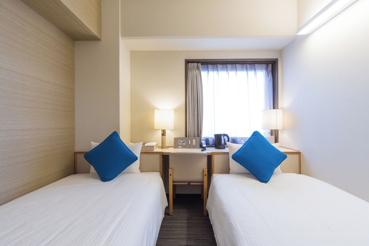 Una habitación de hotel con dos camas individuales, un escritorio, una silla, una lámpara y una ventana con cortinas.