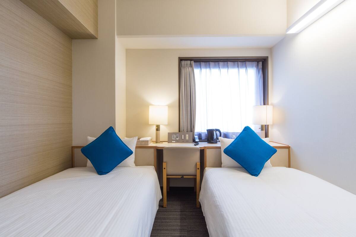 A hotel room with two beds, a desk, a chair, two lamps, a window with curtains, and a coffee machine.