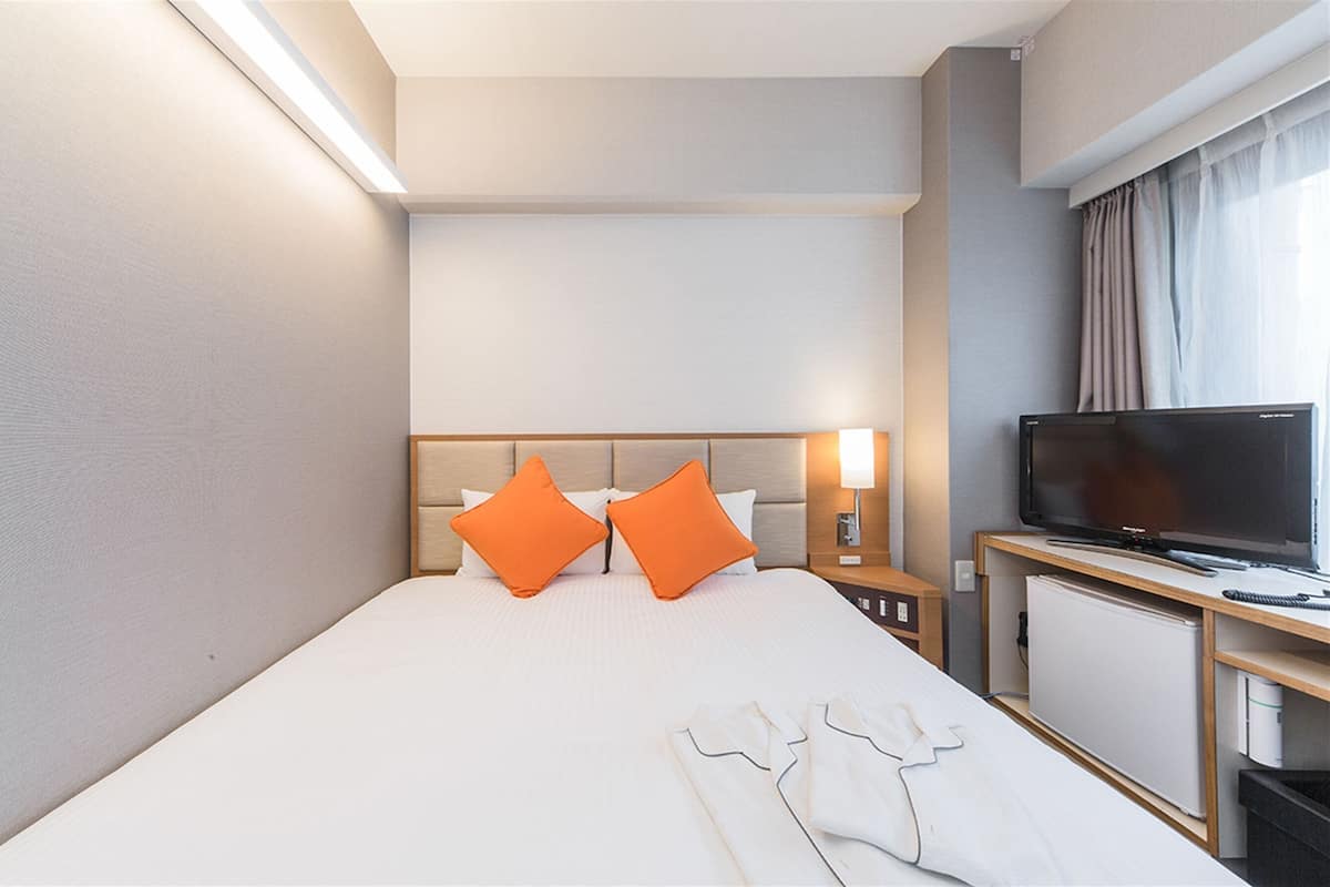 A hotel room with a bed, two orange pillows, a bedside table with a lamp, a TV, and a small refrigerator.