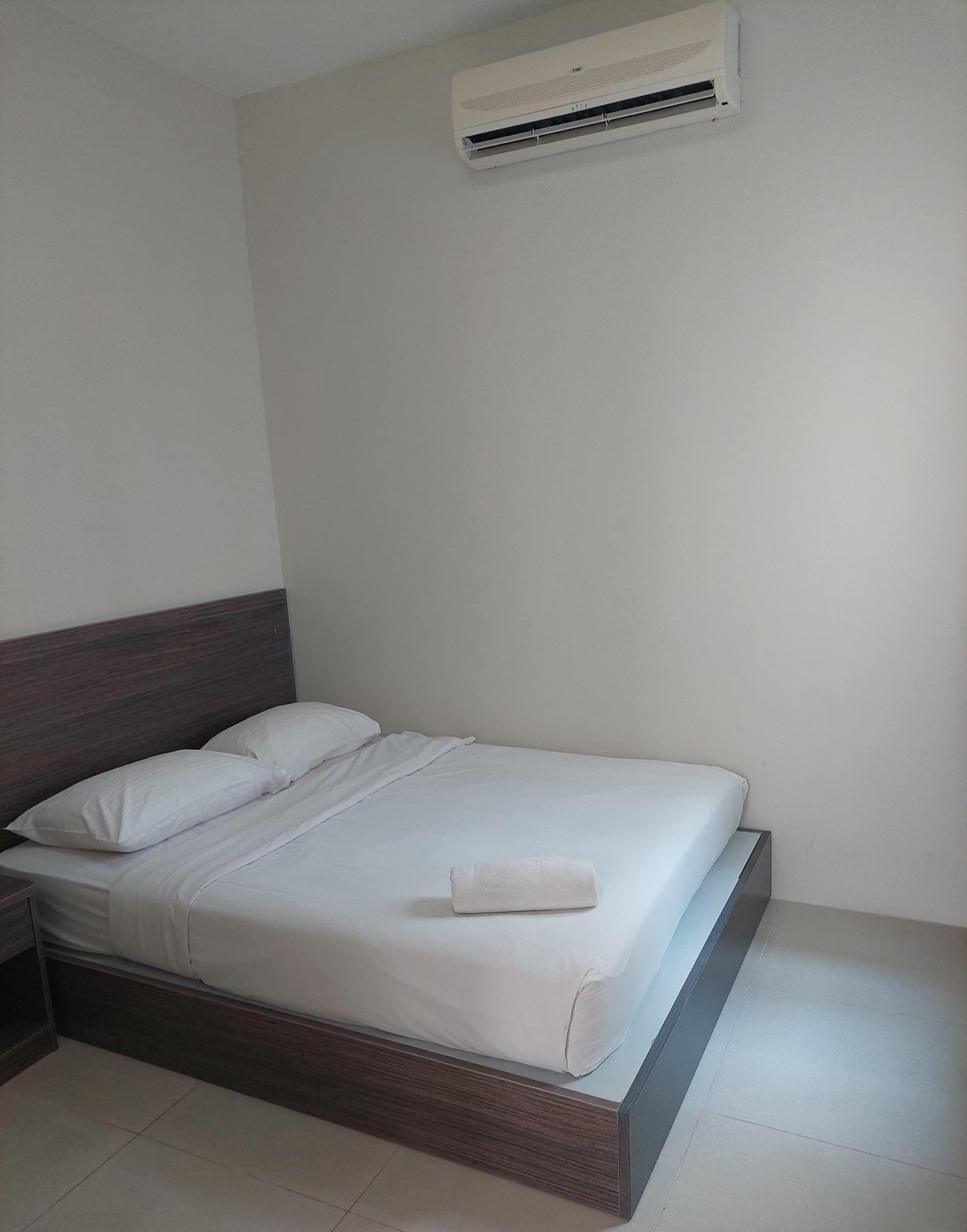 Un lit bien fait, avec du linge de lit blanc et une serviette pliée, une tête de lit en bois et un appareil de climatisation fixé au mur.