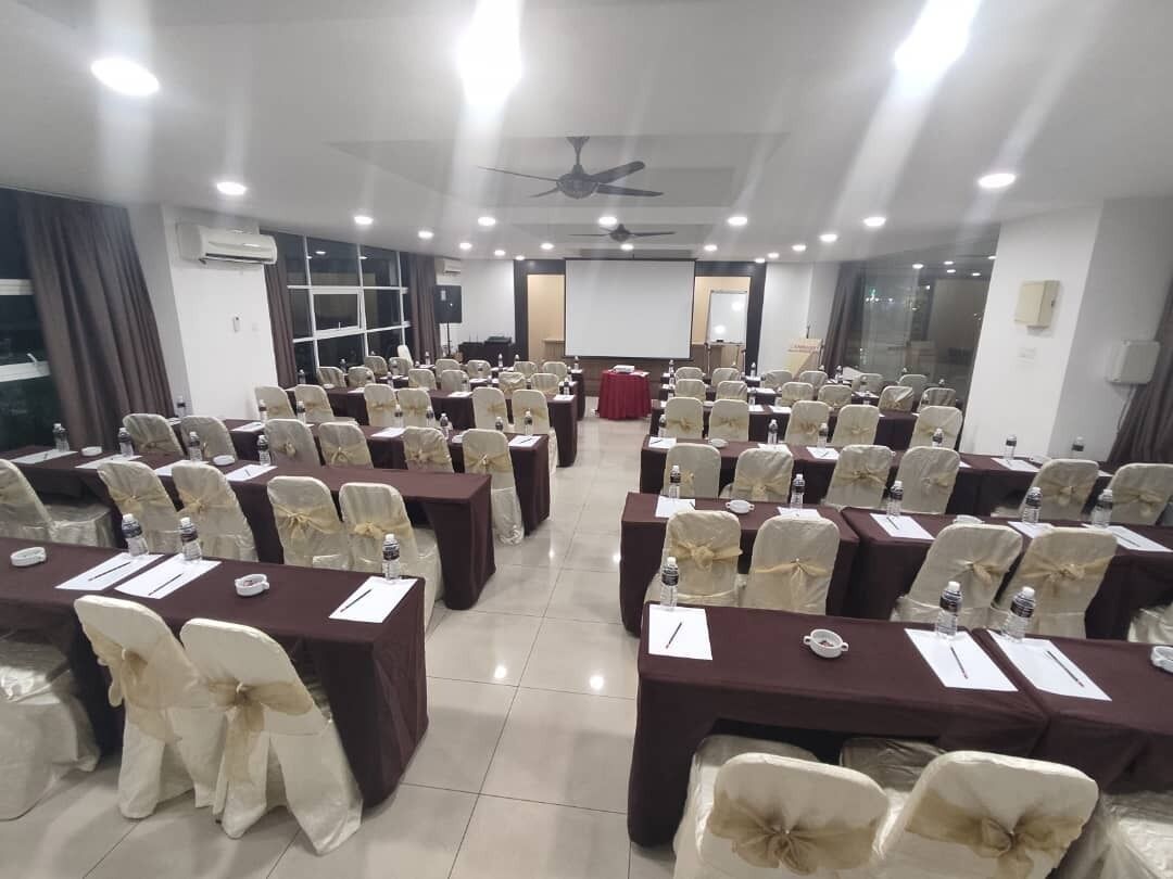 Un salón de conferencias con filas de mesas y sillas, un atril y una pantalla al frente.