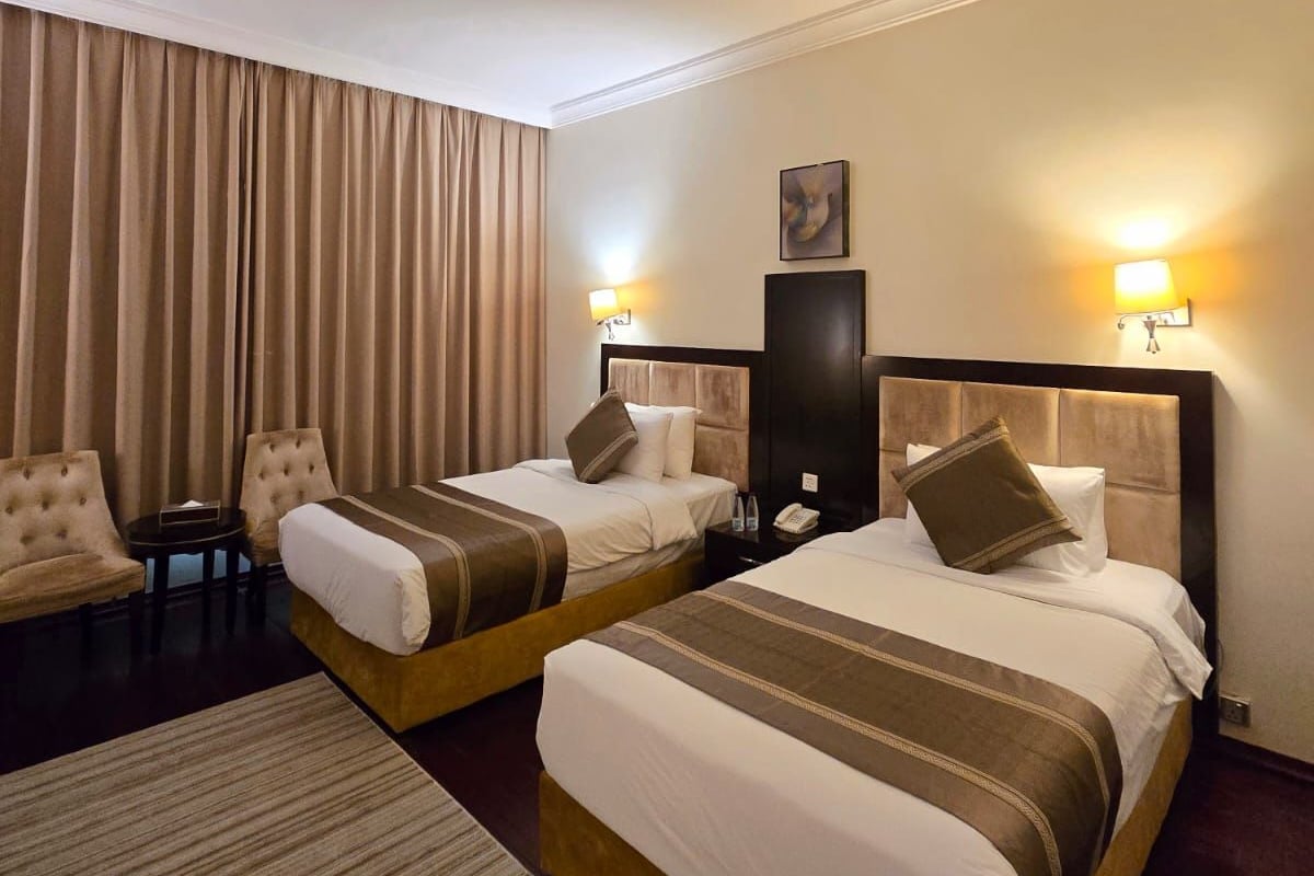 A hotel room with two beds, a desk, chairs, and a large window with curtains.