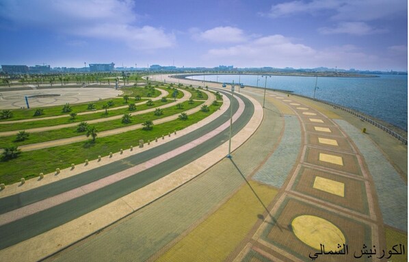 An aerial view of a coastal promenade with curved pathways, green spaces, and a waterfront.