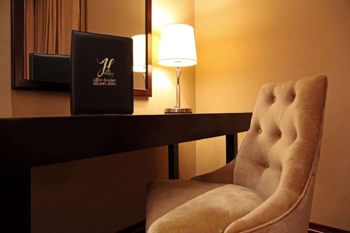 A hotel room with a plush armchair, a bedside table with a lamp, and a framed item with text.