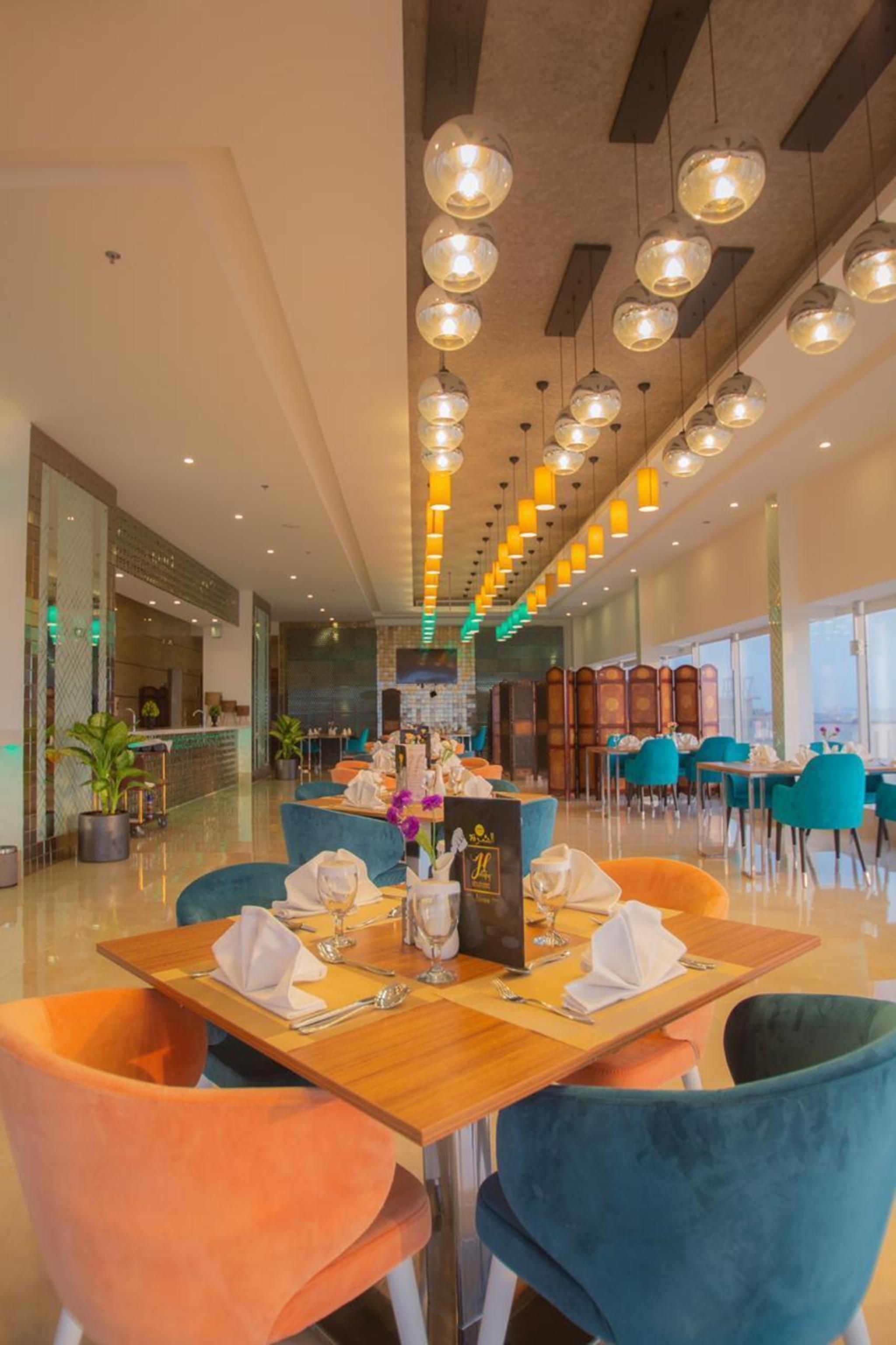 Un comedor moderno con mesas de madera, sillas azul verdosas y anaranjadas, e iluminación colgante.