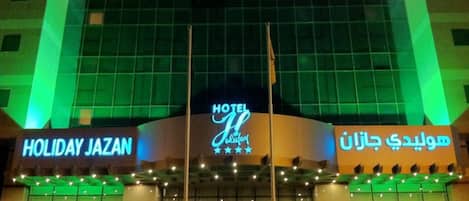 Fachada de hotel con letreros iluminados que dicen "HOLIDAY JAZAN" y "HOTEL JAL" en inglés y árabe, respectivamente.