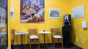 Una habitación pequeña con paredes amarillas, tres sillas de madera y una mesa, una cafetera y tres cuadros en la pared.