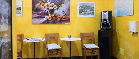 A small, yellow-walled room with three wooden chairs and a table, a coffee machine, and three framed pictures on the wall.