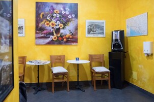 Una habitación pequeña con paredes amarillas, tres sillas de madera y una mesa, una cafetera y tres cuadros en la pared.