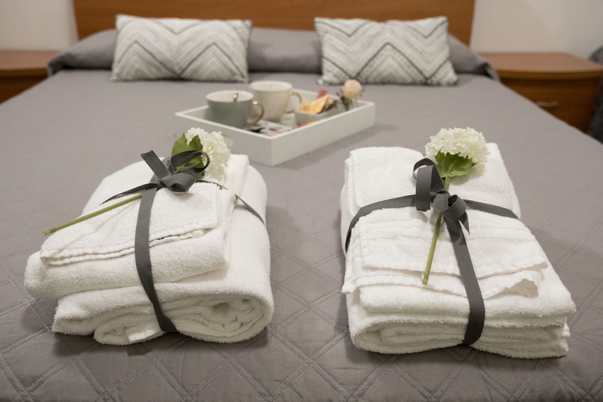 Ein ordentlich bezogenes Bett mit zwei gefalteten Handtüchern, die mit Bändern zusammengebunden sind, einer Ablage mit Tasse und Blumen sowie einem Kissen mit Zickzackmuster.