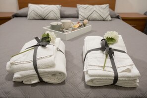 Una cama bien hecha, adornada con dos toallas dobladas sujetas con cintas, una bandeja con una taza y flores, y un cojín con diseño de franjas en zigzag.