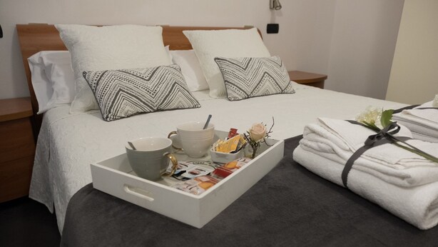 Una cama bien tendida con sábanas blancas, dos almohadas estampadas, una bandeja con una taza, cuencos y revistas, y una pila de toallas dobladas con un lazo.