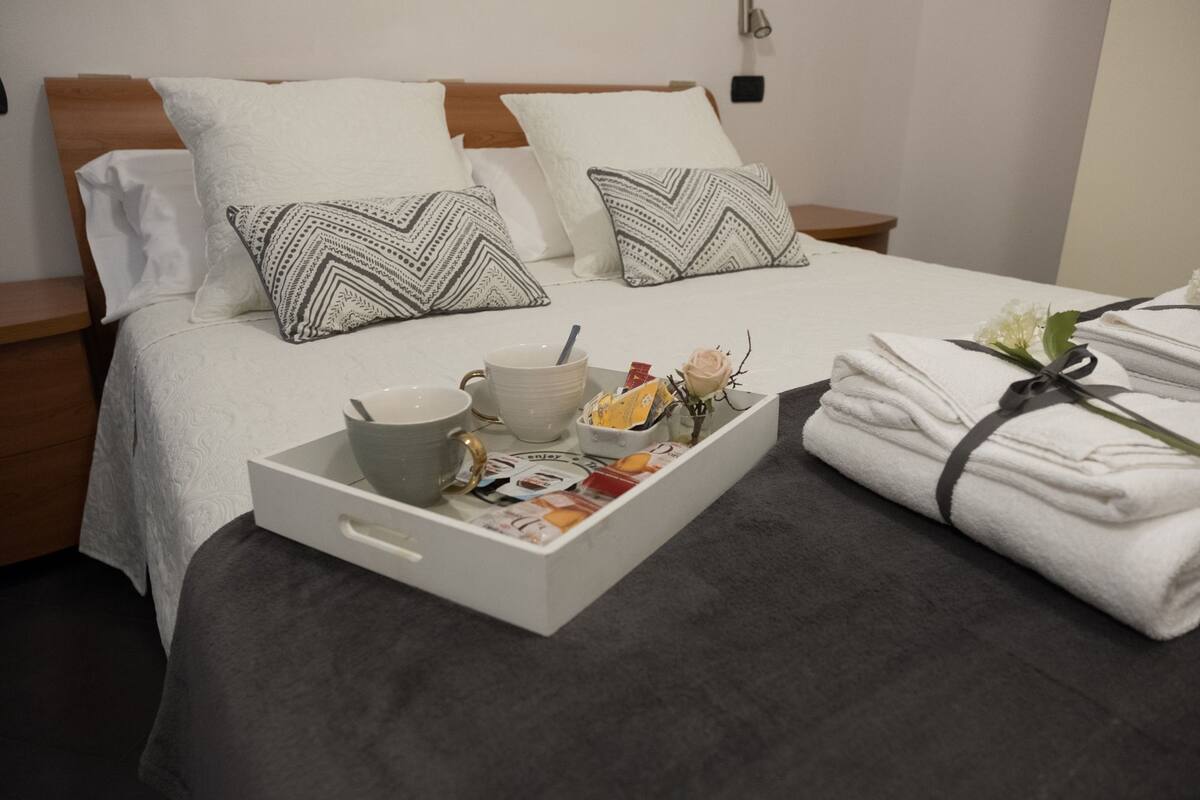 Un lit bien fait, avec des draps blancs, deux coussins à motifs, un plateau avec une tasse, des bols et des magazines, et une pile de serviettes pliées, le tout maintenu par un ruban.