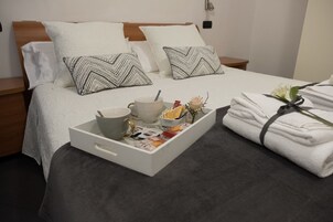 Una cama bien tendida con sábanas blancas, dos almohadas estampadas, una bandeja con una taza, cuencos y revistas, y una pila de toallas dobladas con un lazo.