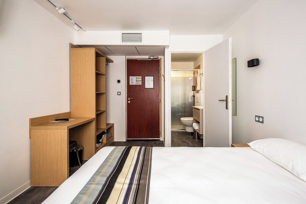 Une chambre d’hôtel avec un lit, un bureau en bois, une armoire et une salle de bain visible à travers une porte ouverte.