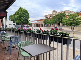 Un área de estar al aire libre con mesas y sillas, un borde decorativo de plantas y vistas a un edificio histórico al fondo.