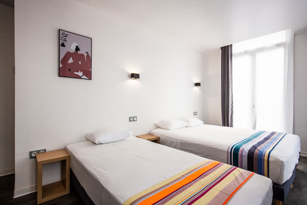 Habitación de hotel con dos camas, una mesita de noche pequeña, una lámpara de pared y una ventana con cortinas.