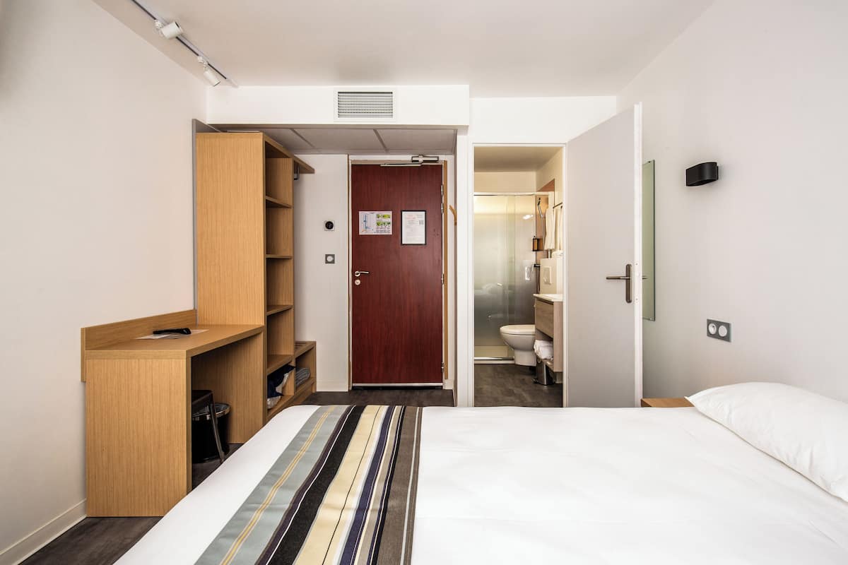 Habitación de hotel con cama, escritorio de madera, armario y baño visible a través de una puerta abierta.