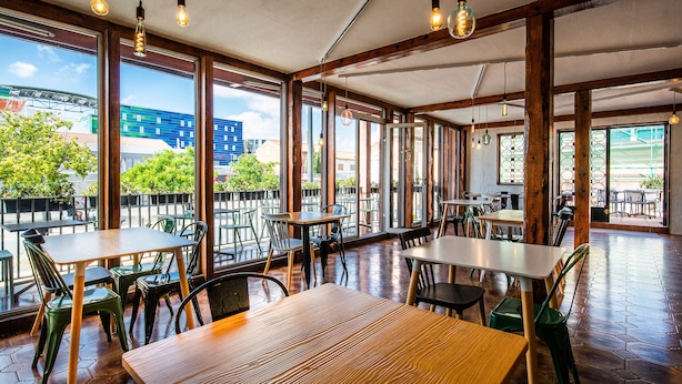 Un comedor interior con mesas y sillas de madera, ventanas amplias y lámparas colgantes.