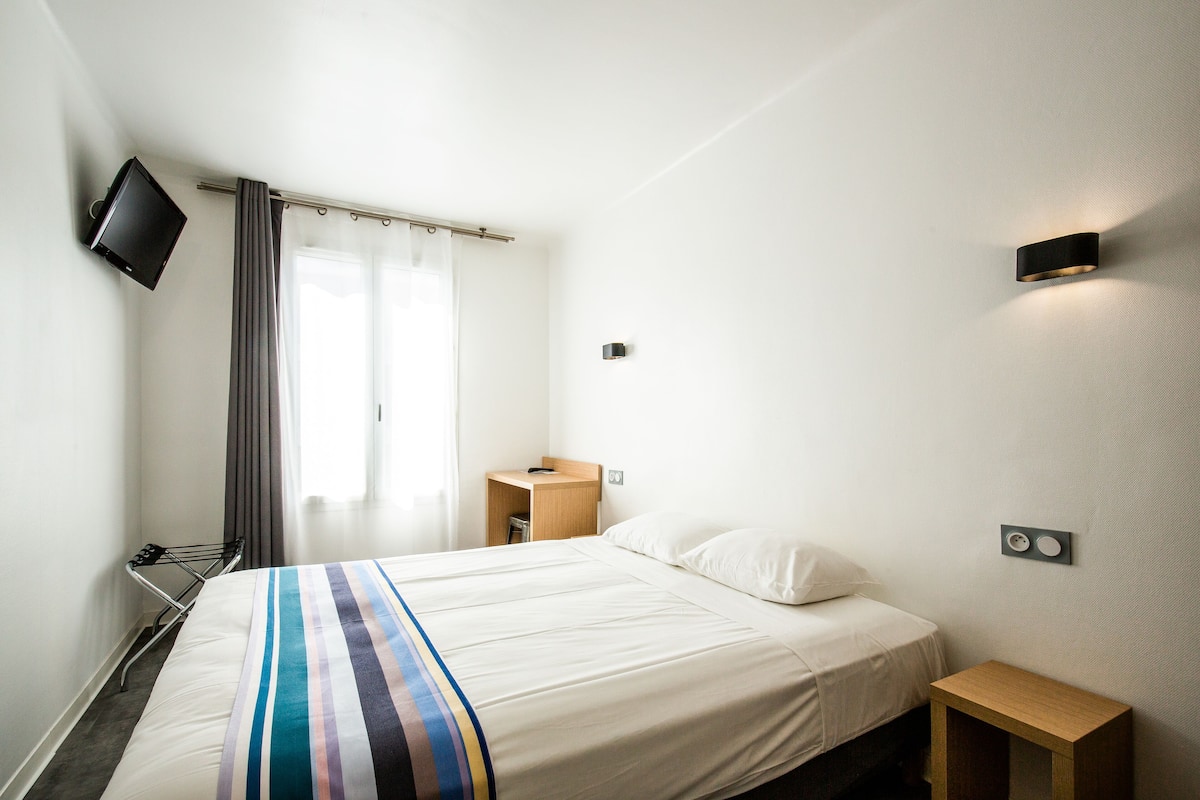 Una habitación de hotel con cama, televisor montado en la pared, una mesita de noche de madera y una ventana con cortinas.