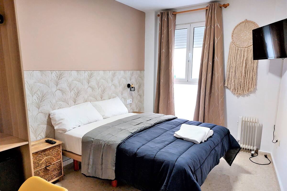 A bedroom with a bed, a bedside table, a chair, a wall-mounted TV, and a window with curtains.