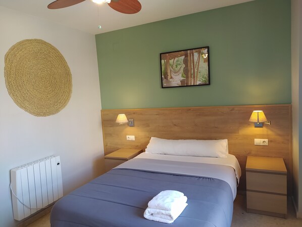 Una cama bien hecha con una toalla doblada, dos lámparas de noche, un cabecero de madera, un cuadro en la pared y un ventilador de techo.