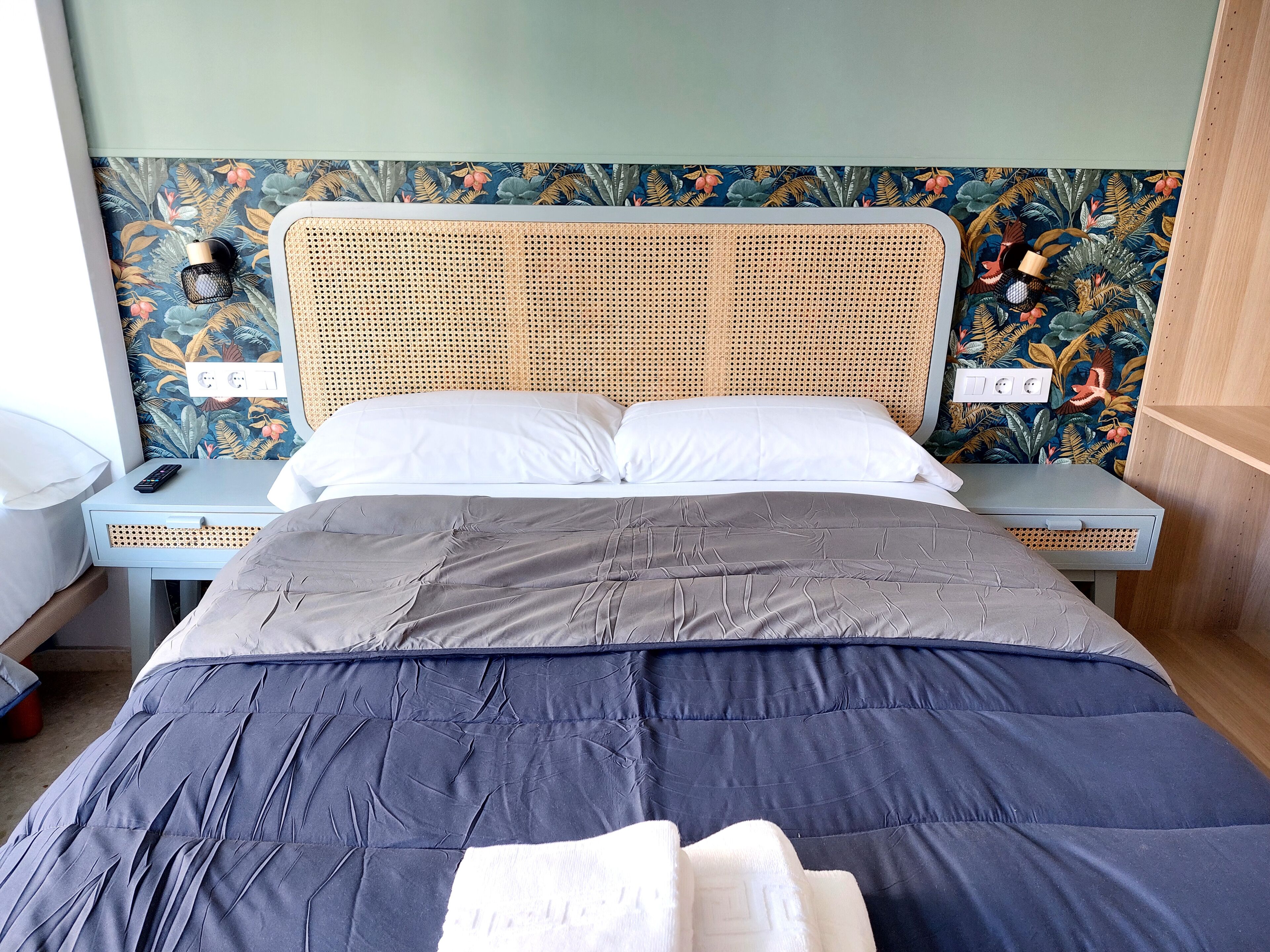 Una cama bien hecha con edredón gris y azul, cabecera de mimbre y un estampado floral en el papel tapiz.