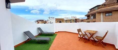A rooftop terrace with a green artificial turf area, two lounge chairs, and a wooden table with chairs.