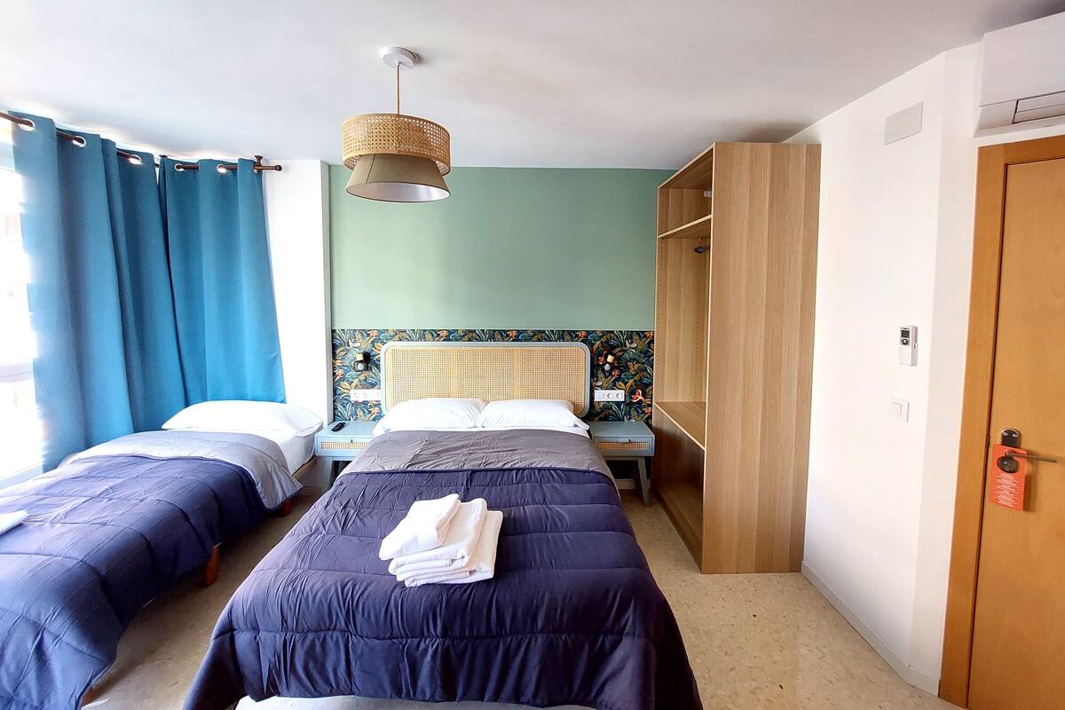 A hotel room with two beds, a large window with curtains, a woven ceiling lamp, a wooden wardrobe, and a wall-mounted thermostat.