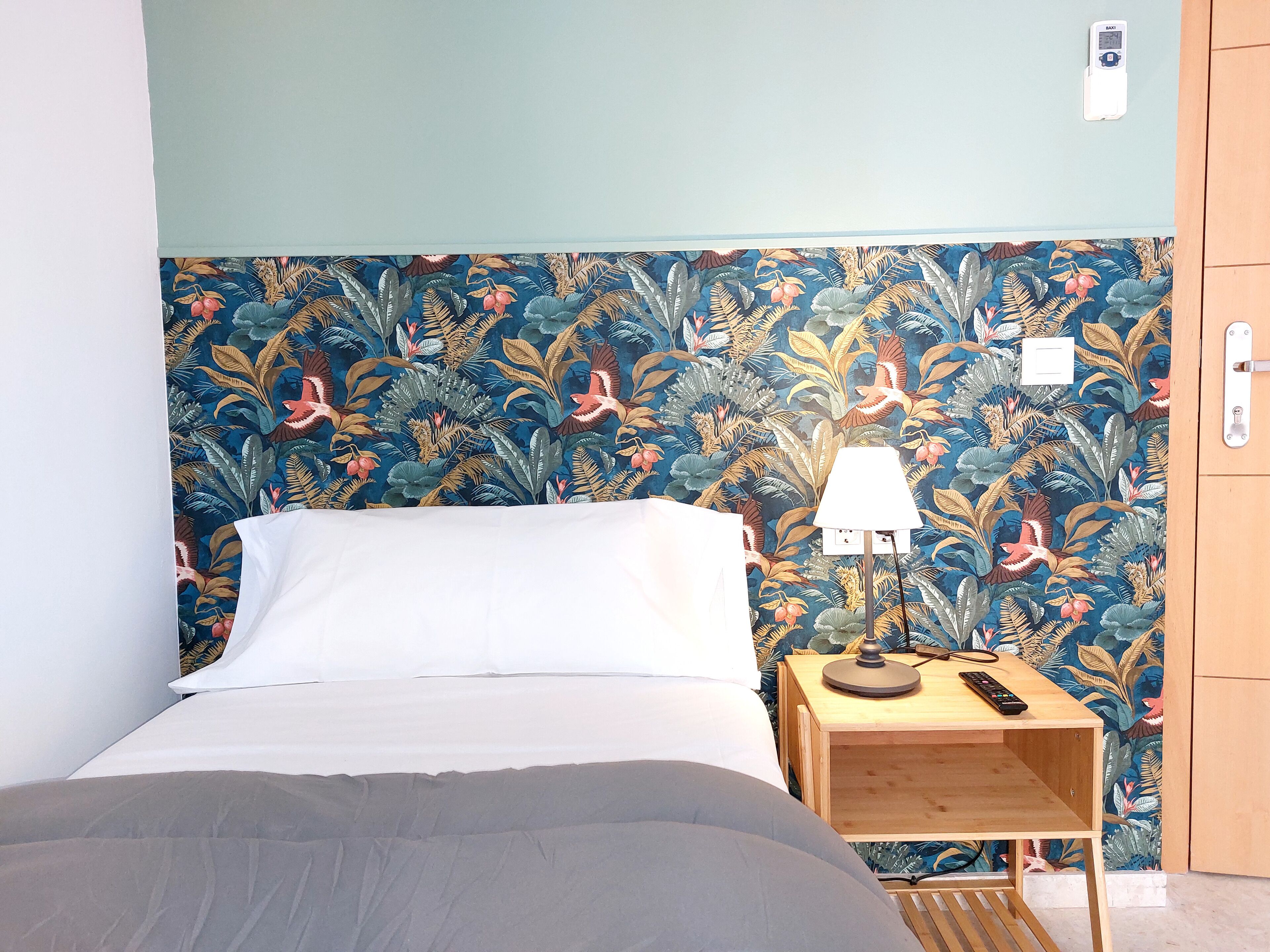 Un dormitorio con una cama, una mesita de noche con una lámpara y un papel tapiz tropical en la pared.