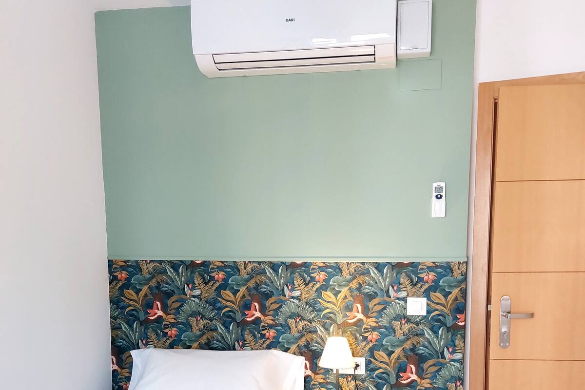 A bedroom with a bed, a nightstand with a lamp, an air conditioning unit, and a wooden door.