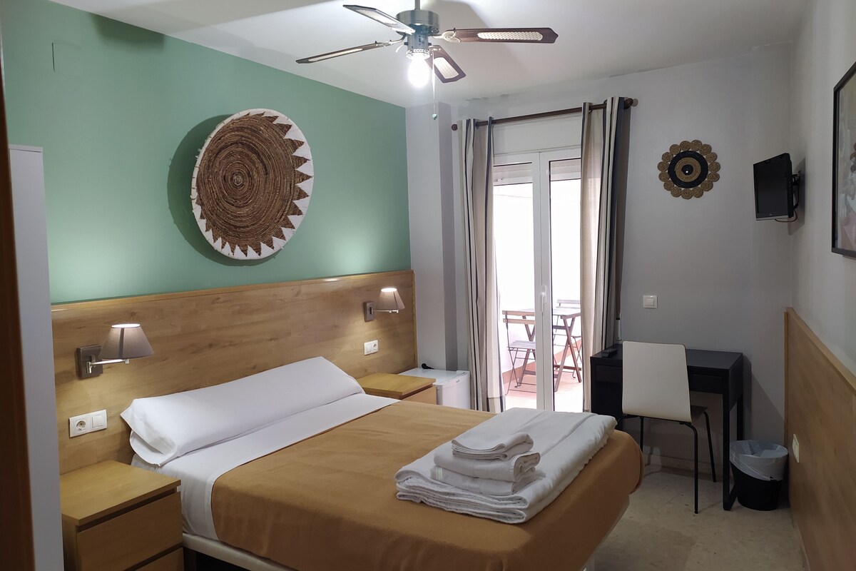 A hotel room with a bed, a desk, a chair, a ceiling fan, and a view of the outside.