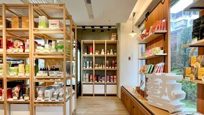 Interior de una tienda con estantes de madera llenos de diversos productos, un mostrador con artículos y vistas a la vegetación exterior.