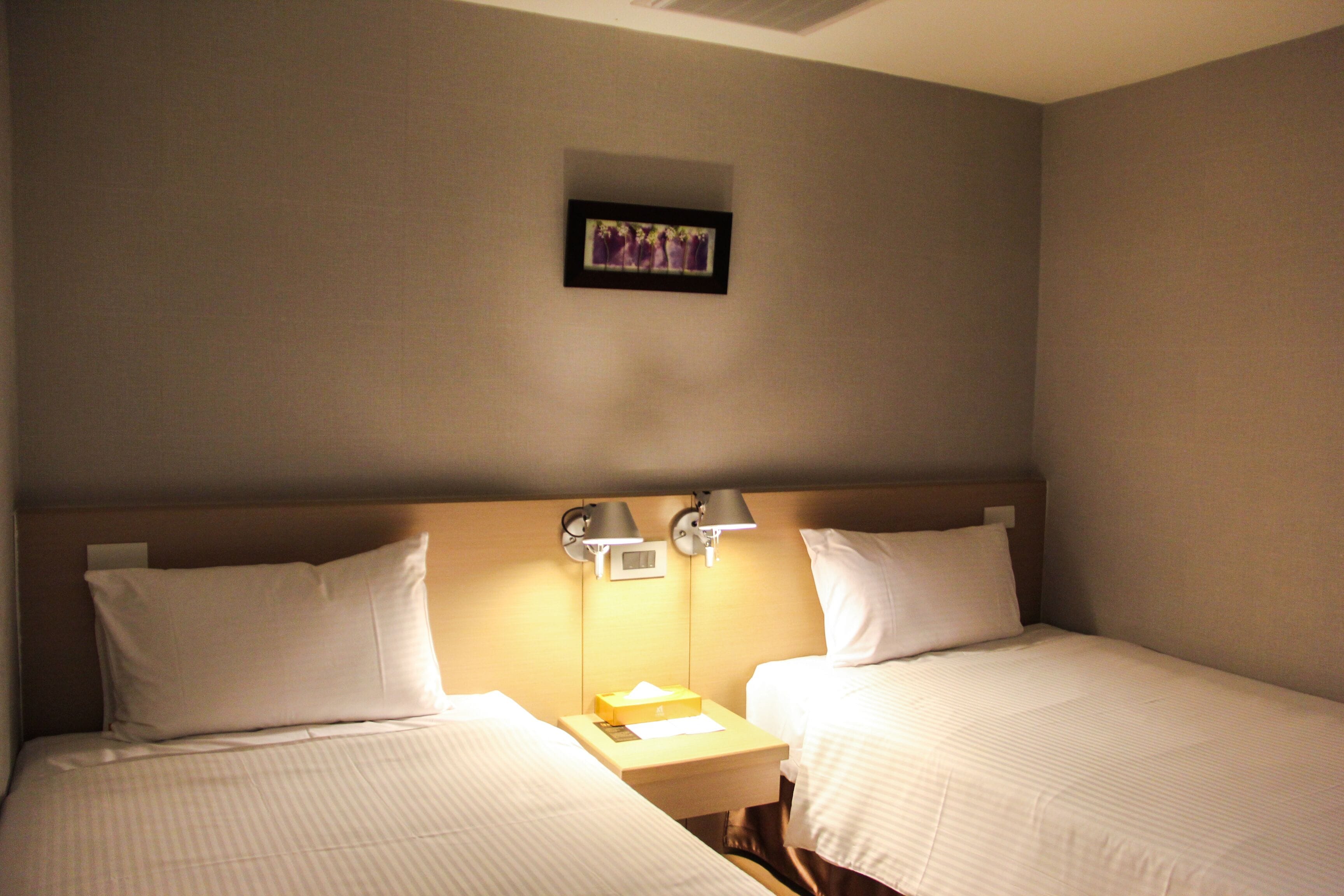 A hotel room with two single beds, a nightstand with a lamp, and a framed picture on the wall.