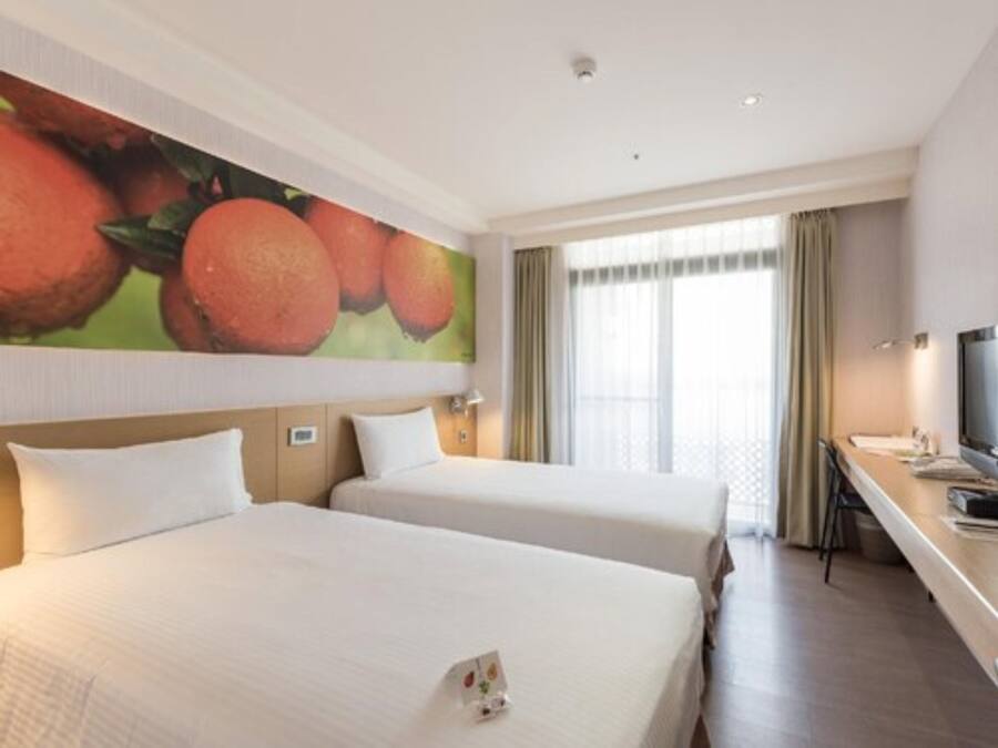 Camera d'albergo con due letti, un quadro di grandi dimensioni a tema frutta, una scrivania con televisore e una finestra con tende.