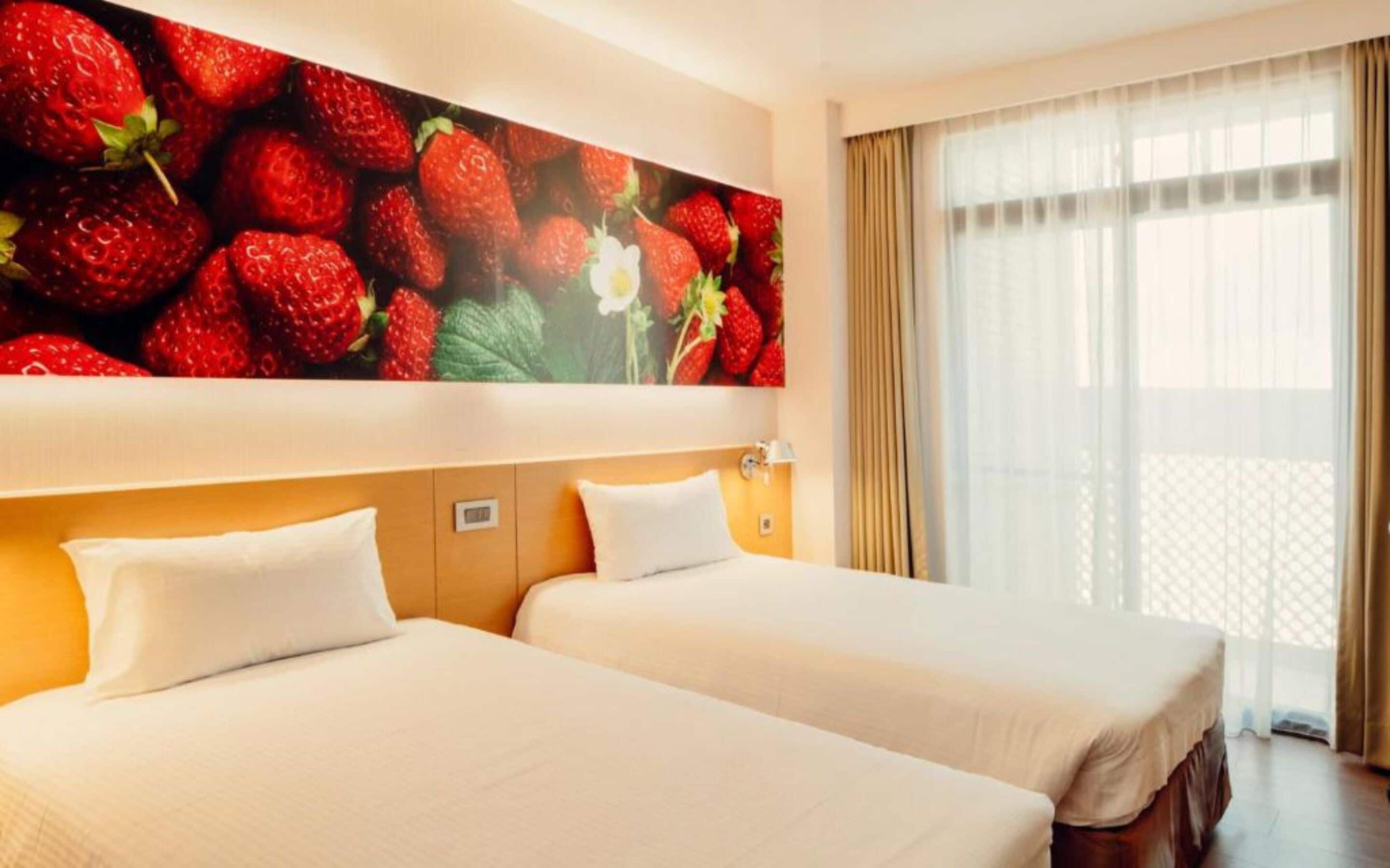 A hotel room with two beds, a large wall mural of strawberries, a window with curtains, and a visible thermostat.