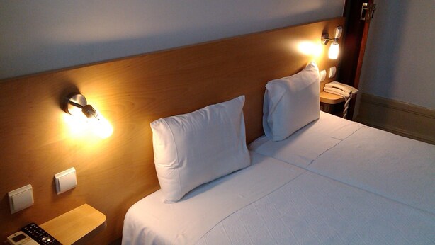Une chambre d’hôtel avec une tête de lit en bois, deux oreillers blancs, une table de chevet avec un téléphone et une télécommande, et un luminaire fixé au mur.
