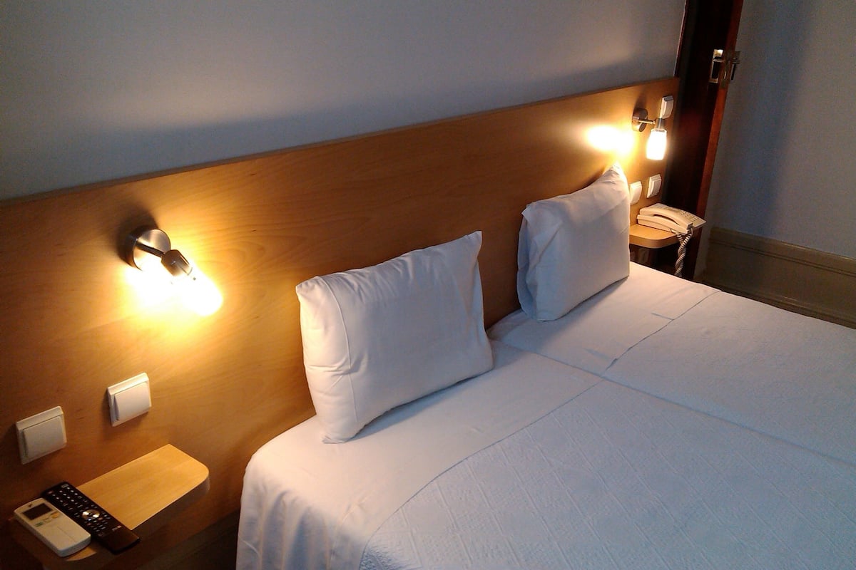 A hotel room with a wooden headboard, two white pillows, a bedside table with a phone and a remote control, and a wall-mounted light fixture.