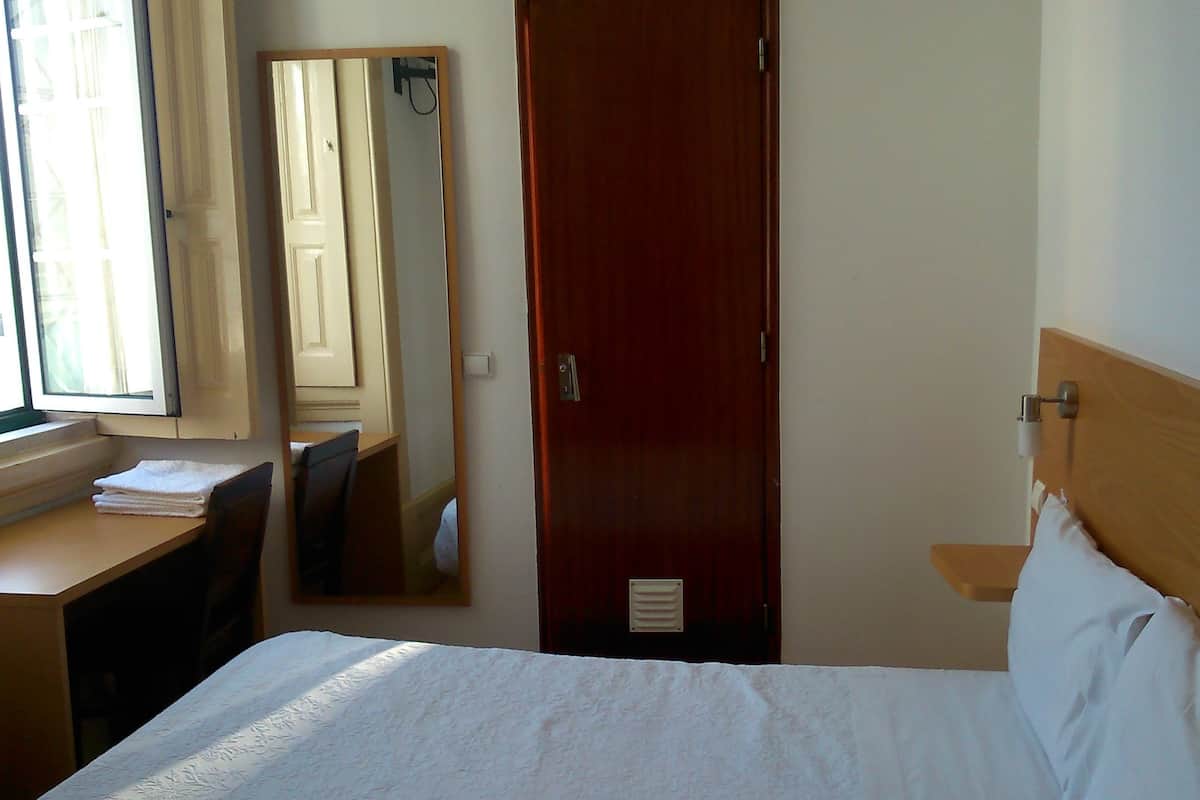 A hotel room with a bed, a desk, a chair, a window, and a door.