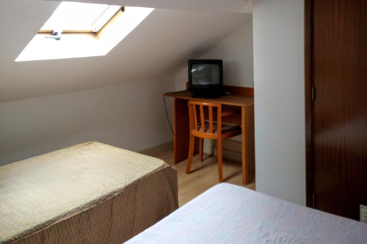 A small, dimly lit room with a single bed, a desk with a computer, and a skylight.