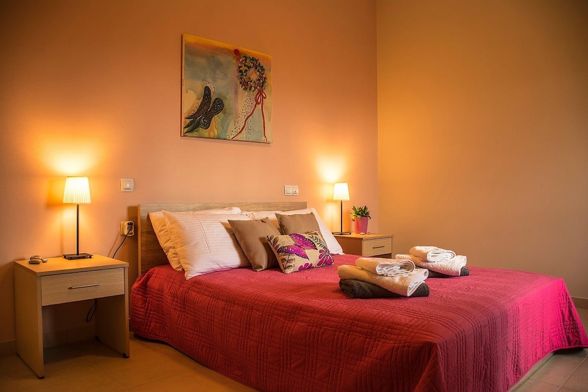 Una cama bien hecha con colcha roja, dos lámparas de noche, una mesita de noche con lámpara, un cuadro en la pared y una pequeña planta en maceta.