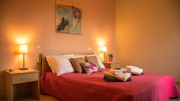 A neatly made bed with a red bedspread, two bedside lamps, a nightstand with a lamp, a wall painting, and a small potted plant.