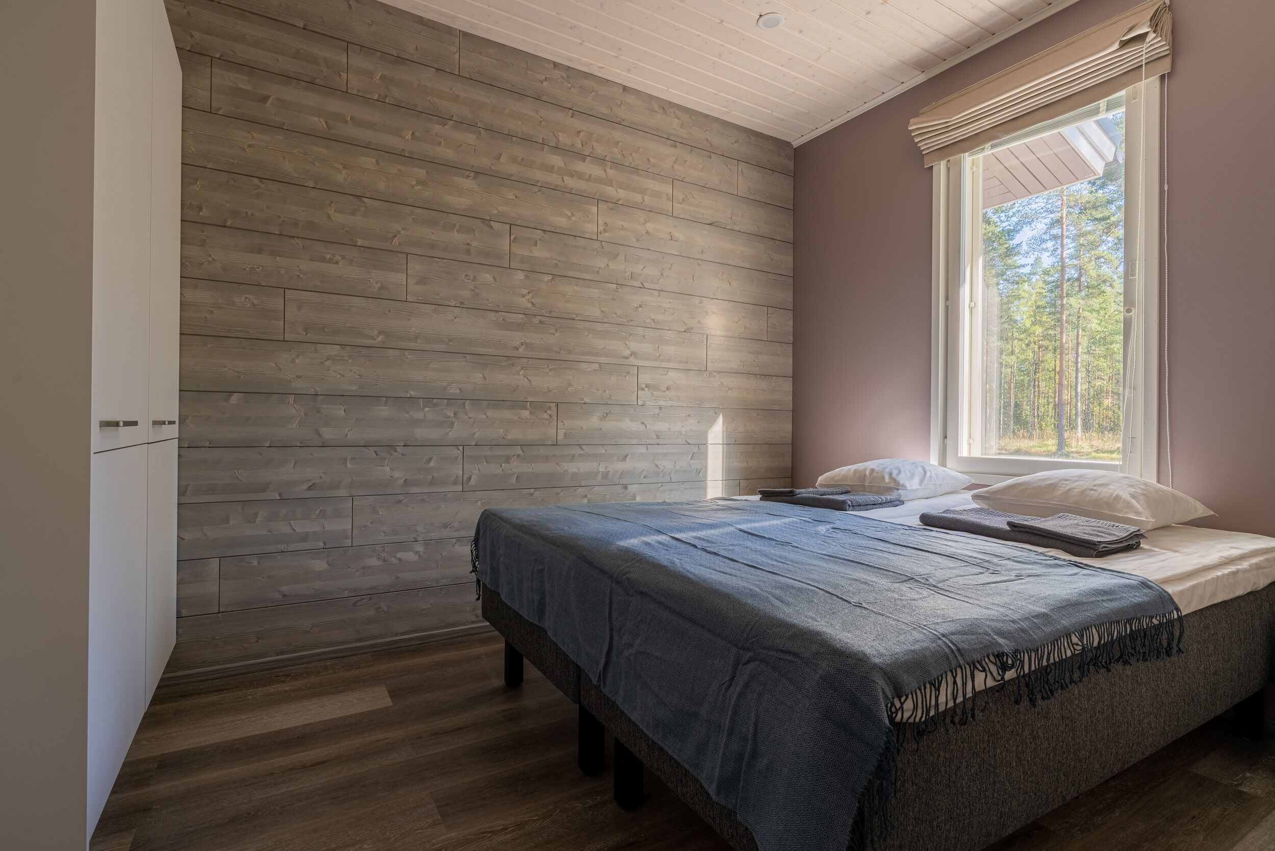Quarto com parede de madeira, janela com persianas, cama com colcha azul e mesa de cabeceira.