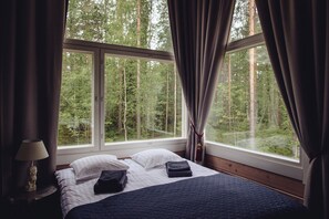 A bedroom with a large window, a bed with white and dark blue bedding, and a lamp on a bedside table.