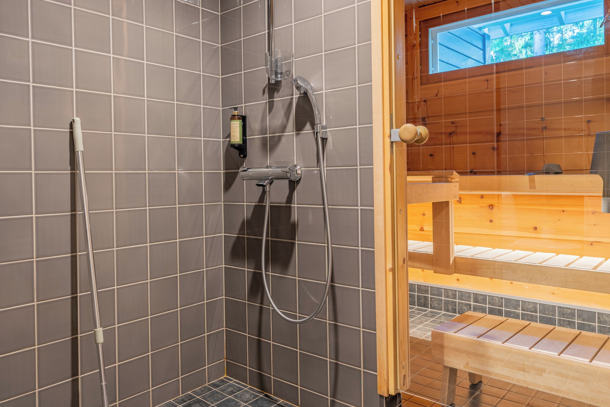 A shower area with tiled walls, a handheld showerhead, a bottle of shampoo, and a wooden bench.
