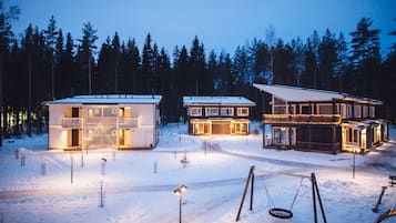 Paisagem coberta por neve com três edifícios, cada um com janelas iluminadas e telhado com neve.